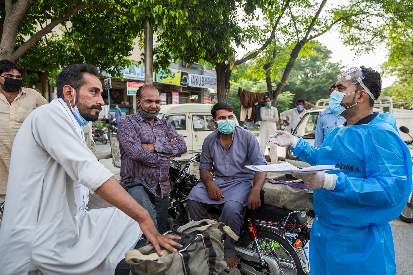 Dr Muhammad Asif is stood on the street interviewing four wage workers sat on and around their motorcycles.  